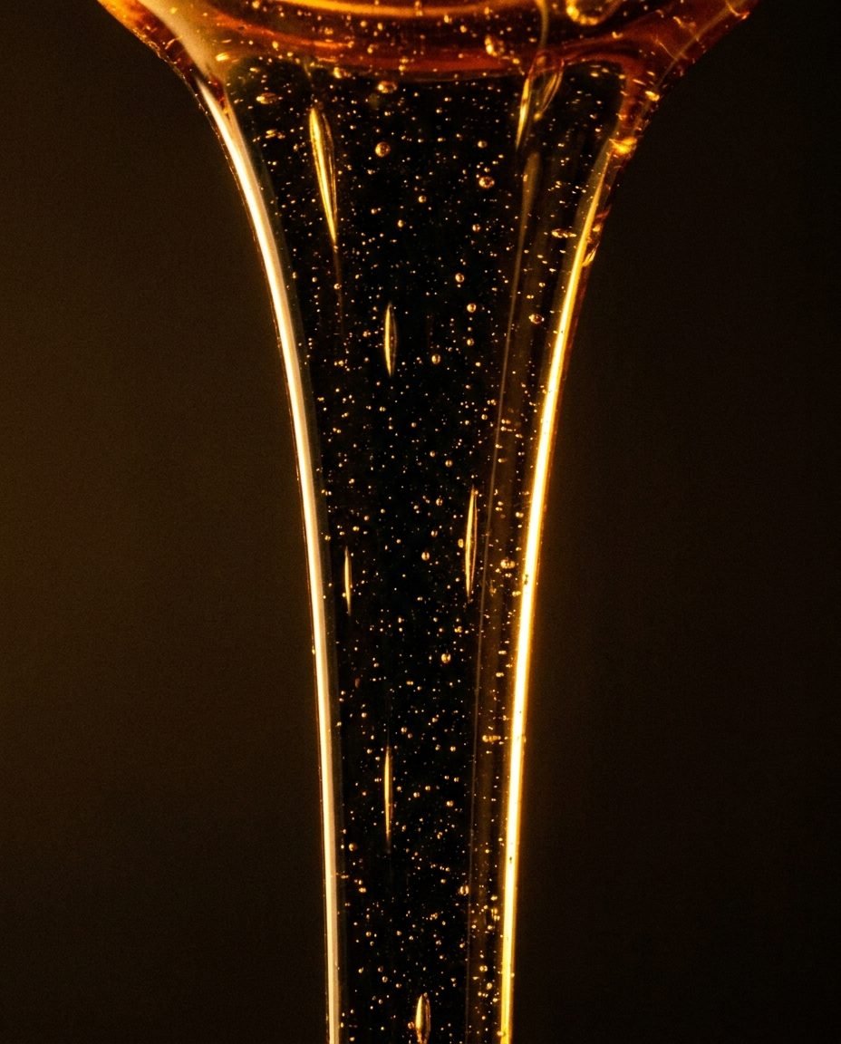 A thin ribbon of very dark amber honey pouring against a black background, backlit with golden light